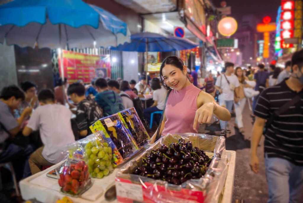 woman standing beside a fruit stall at night in Sampheng Market Bangkok surrounded by colorful produce
