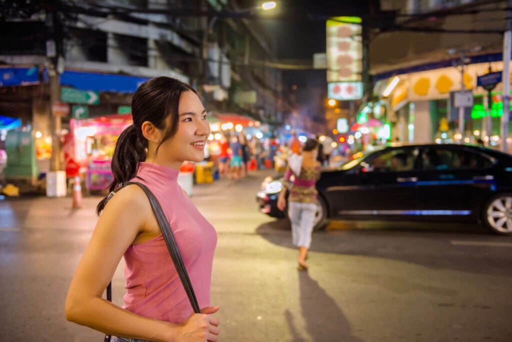woman standing on a busy main road in Chinatown Bangkok at night beside street food stalls and glowing shop signs