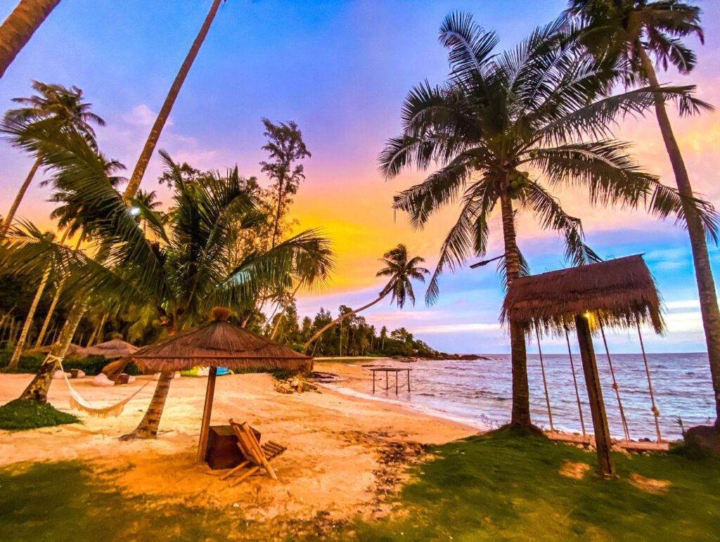 Evening view of a stunning Koh Kood beach with straw huts, swings by the sea, and a vibrant sunset sky.
