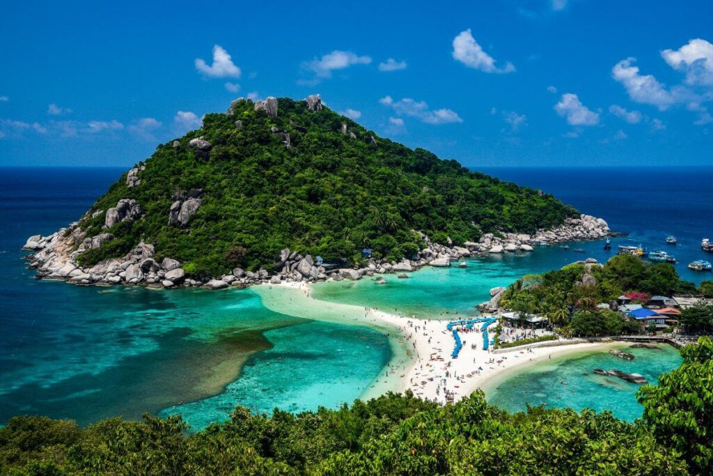 Aerial view of Koh Tao bay with vibrant blue waters, surrounded by lush greenery, seen from a cliff under a clear sky.