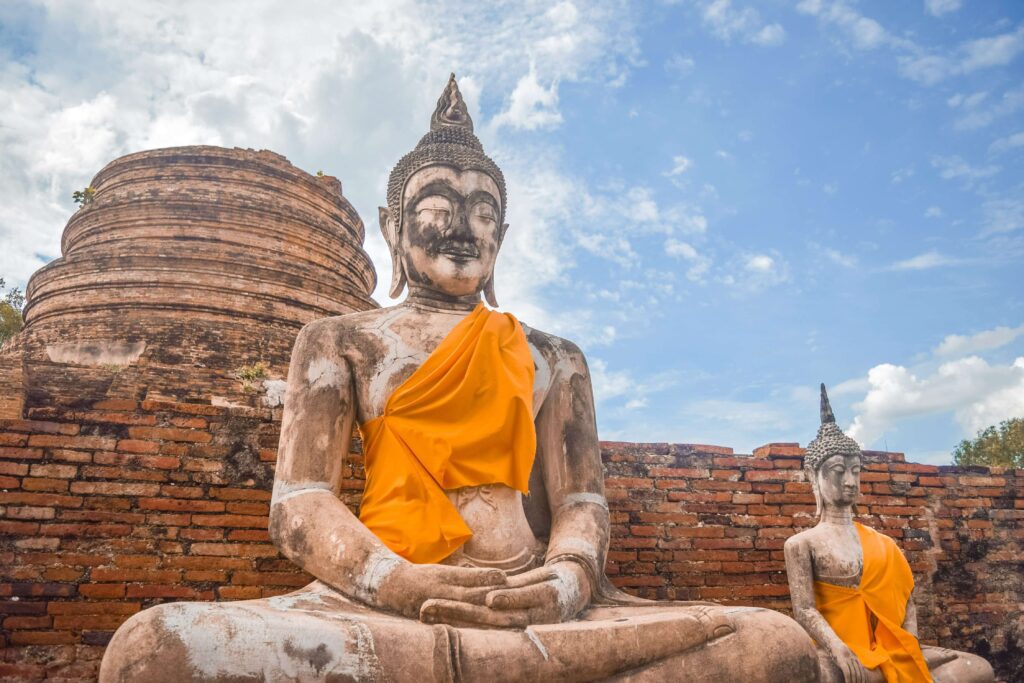 A large, seated stone Buddha statue, weathered and worn, situated among the ancient brick temple ruins in Ayutthaya Historical Park.